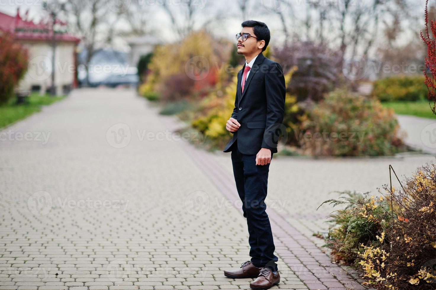 indischer junger mann mit brille, trägt einen schwarzen anzug mit roter krawatte im freien. foto