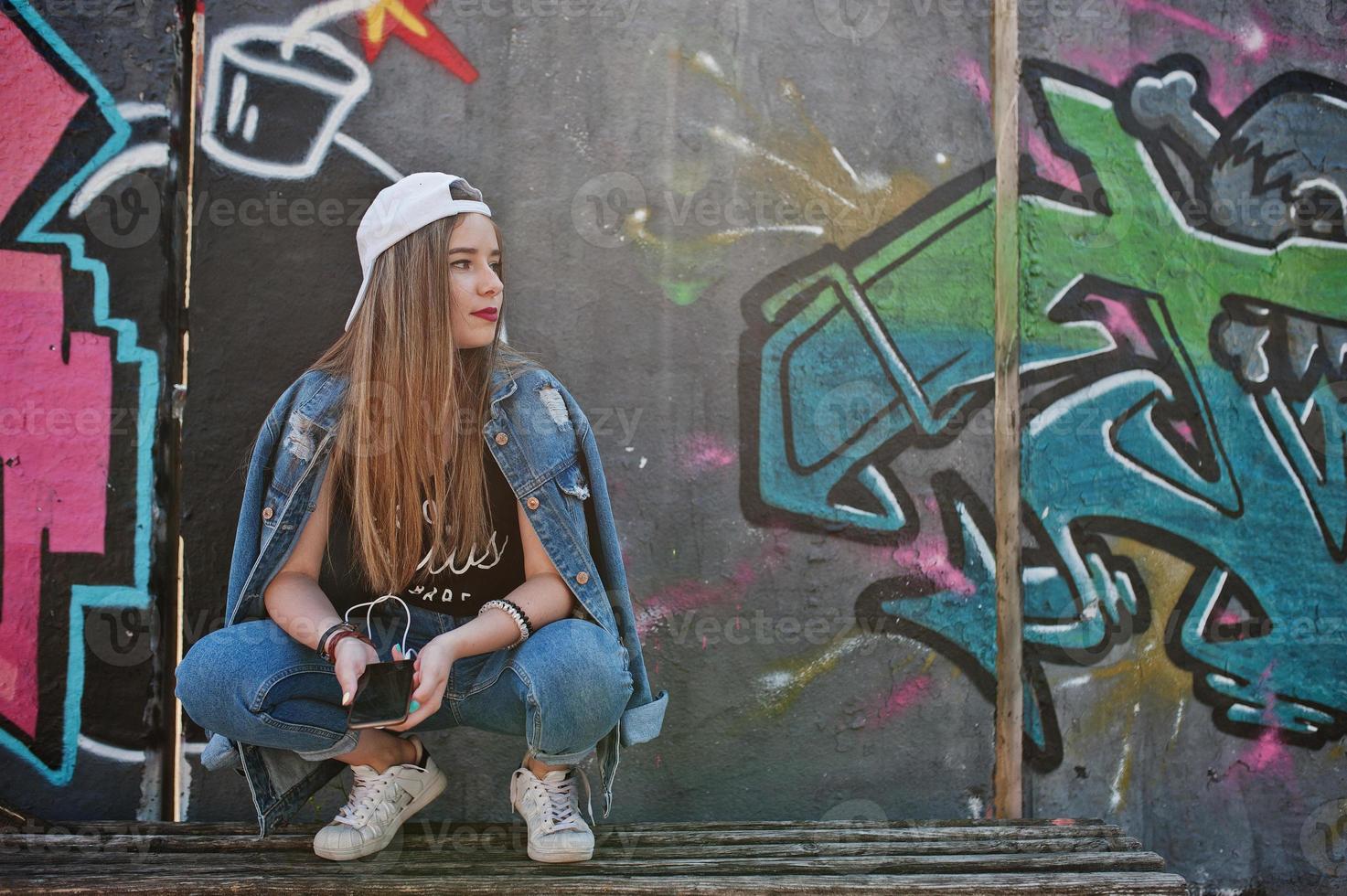 stilvolles, lässiges hipster-mädchen in mütze und jeans, das musik über kopfhörer des handys gegen große graffitiwand mit bombe hört. foto