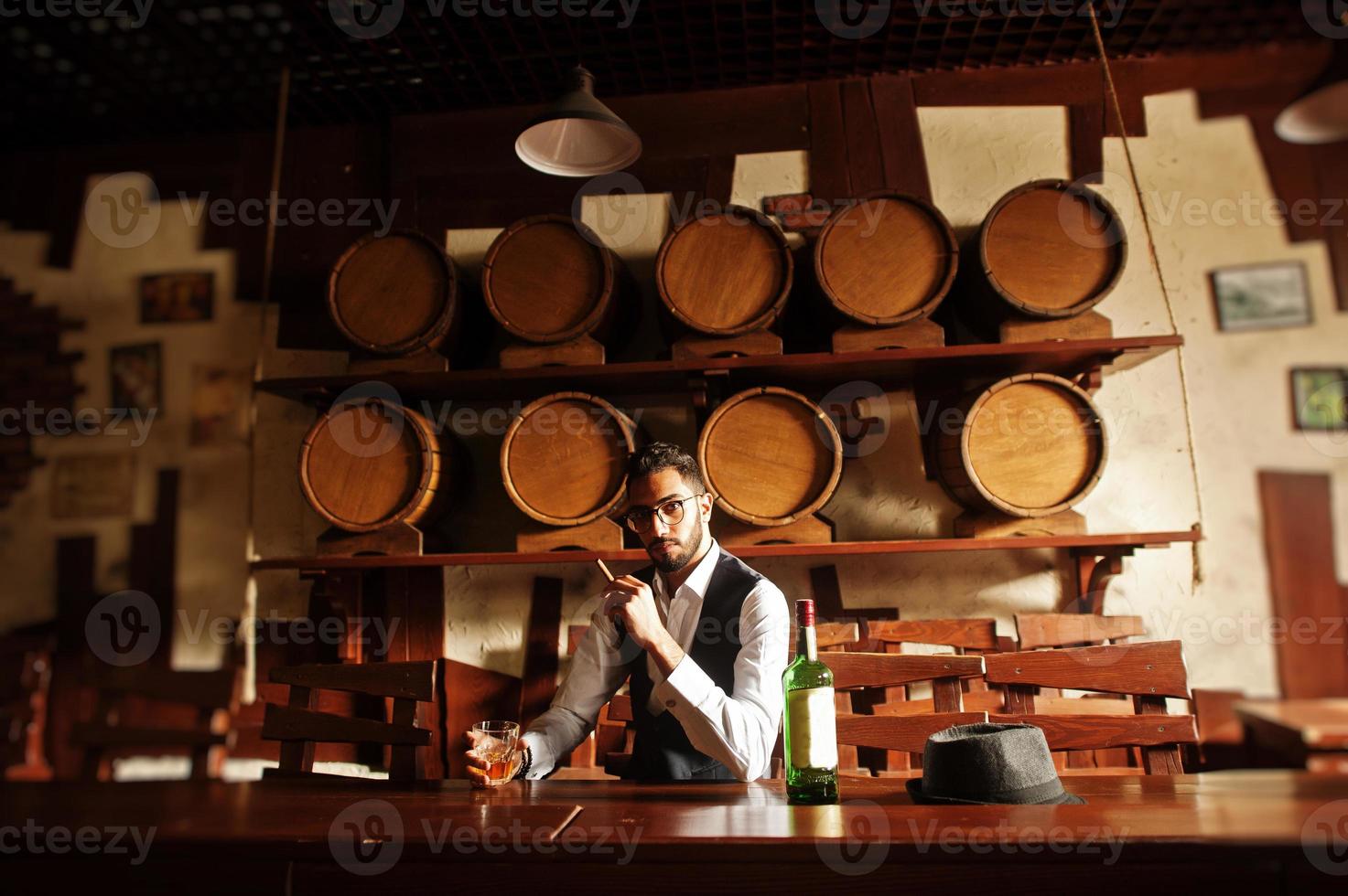 hübscher, gut gekleideter arabischer mann mit einem glas whisky und einer zigarre, die im pub posiert. foto