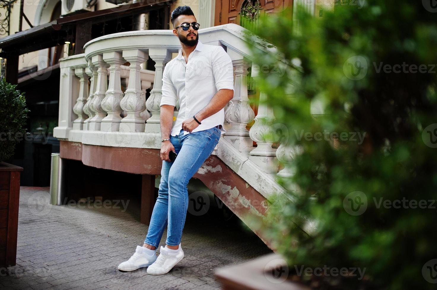 Stilvolles großes arabisches Mannmodell in weißem Hemd, Jeans und Sonnenbrille posierte auf der Straße der Stadt. Bart attraktiver arabischer Typ. foto