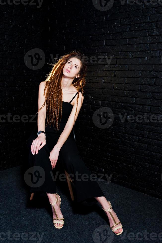 Studioaufnahme eines Mädchens in Schwarz auf einem Stuhl mit Dreadlocks auf Backsteinhintergrund. foto