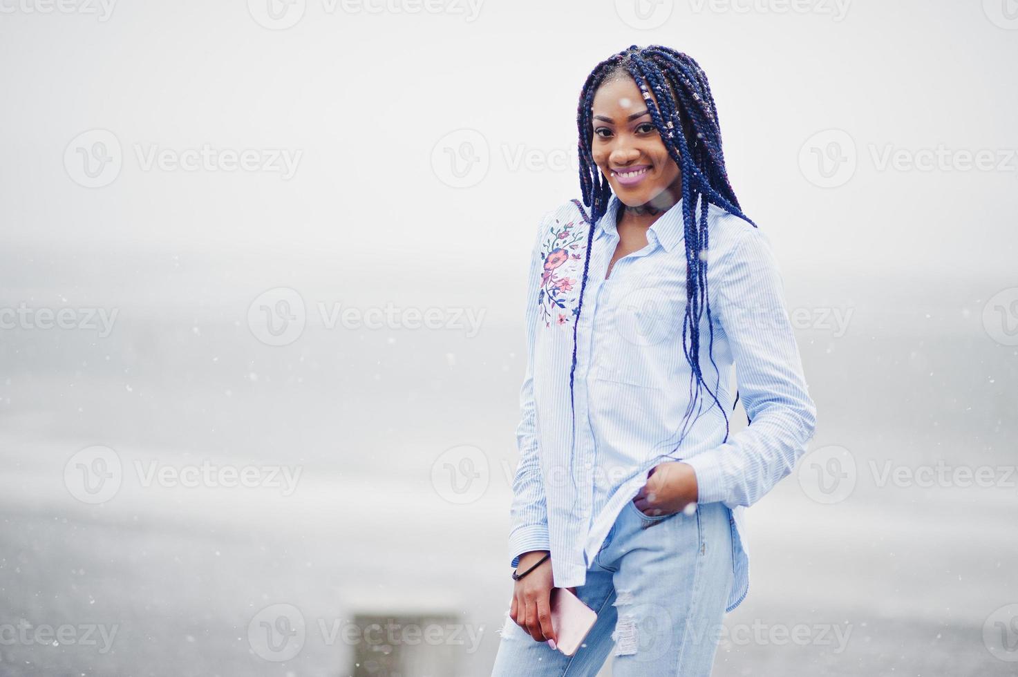 Stilvolles afrikanisch-amerikanisches Mädchen mit Dreadlocks, das Handy zur Hand hält, im Freien auf Pier gegen zugefrorenen See bei Schneewetter. foto