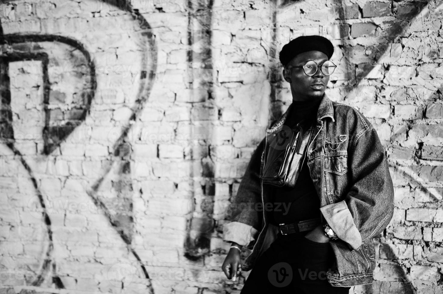 afroamerikanischer mann in jeansjacke, baskenmütze und brille gegen graffiti-ziegelwand mit bruk-schild. foto