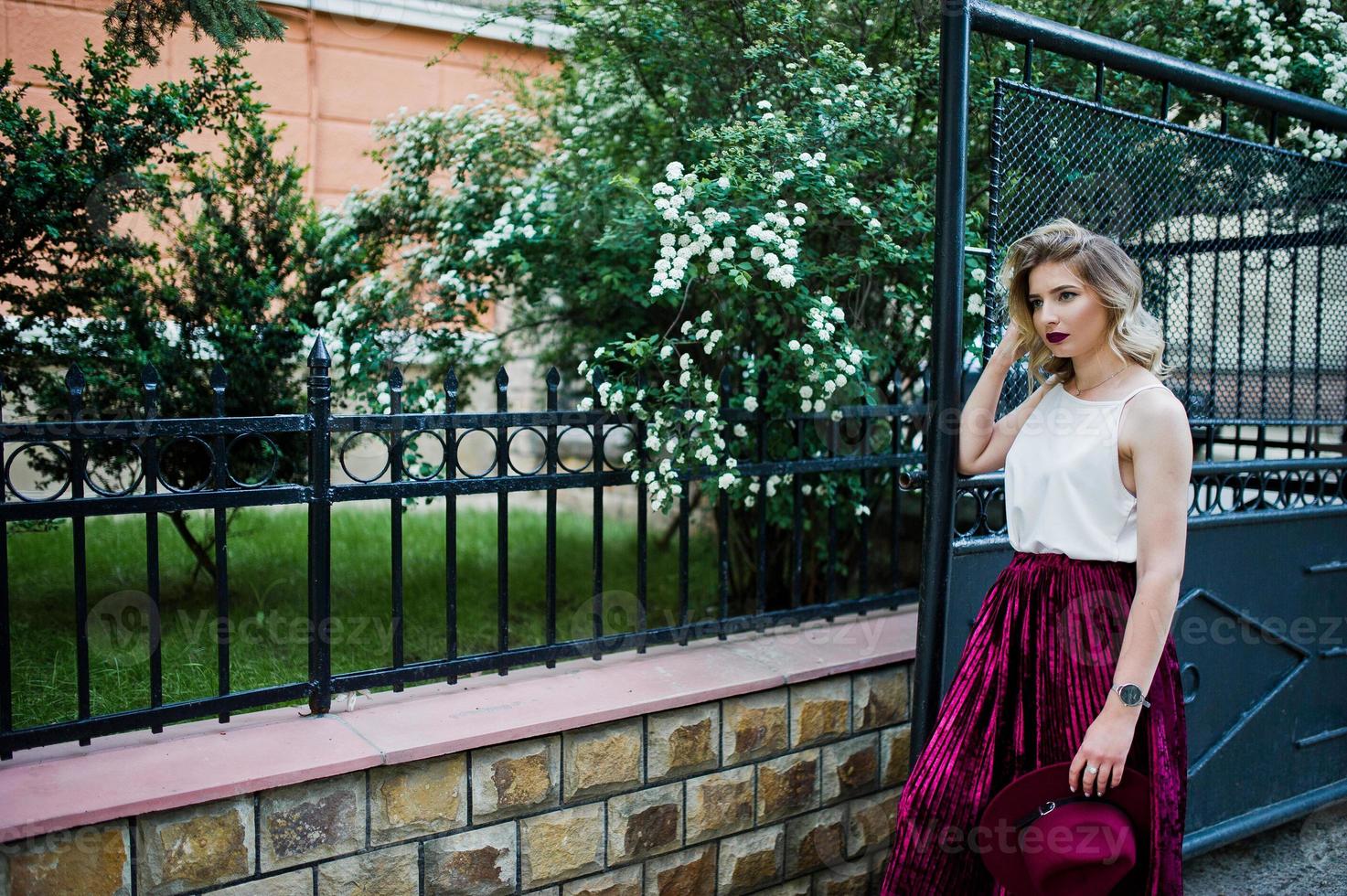 Modisches und schönes blondes Modelmädchen in stylischem rotem Samt-Veloursrock, weißer Bluse und Hut, posiert im Freien vor Toren. foto