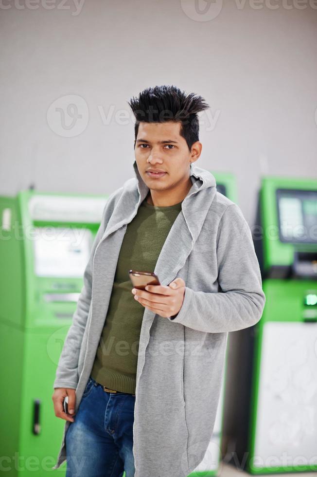 junger stilvoller asiatischer mann mit handy gegen reihe von grünen geldautomaten. foto