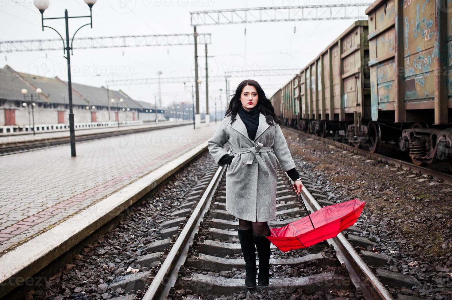 Brünettes Mädchen im grauen Mantel mit rotem Regenschirm im Bahnhof. foto