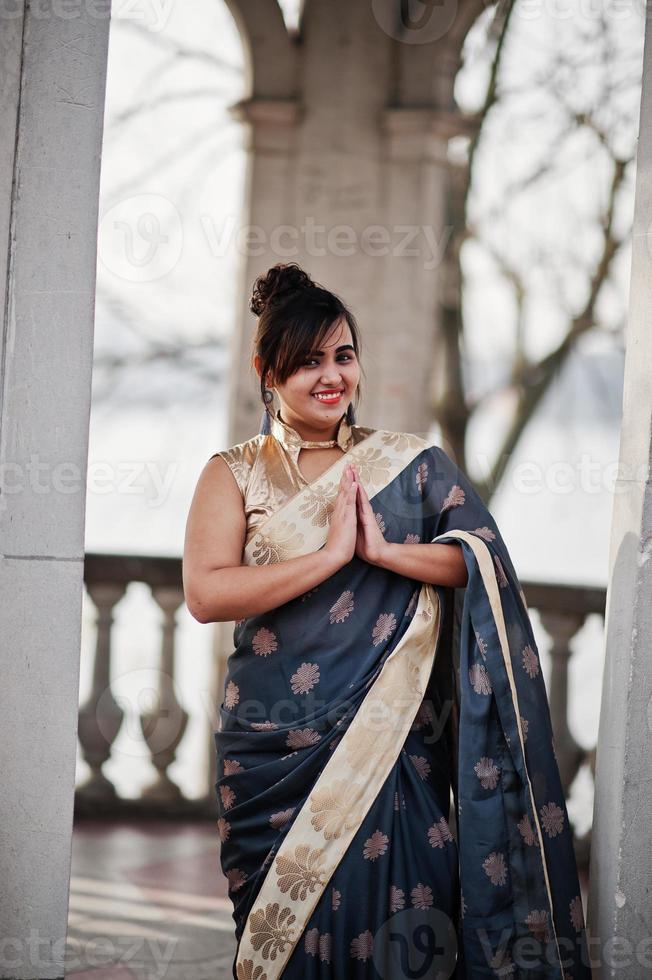 elegantes brünettes südasiatisches indisches mädchen im saree, das unter bogen steht und namaste-handzeichen zeigt. foto