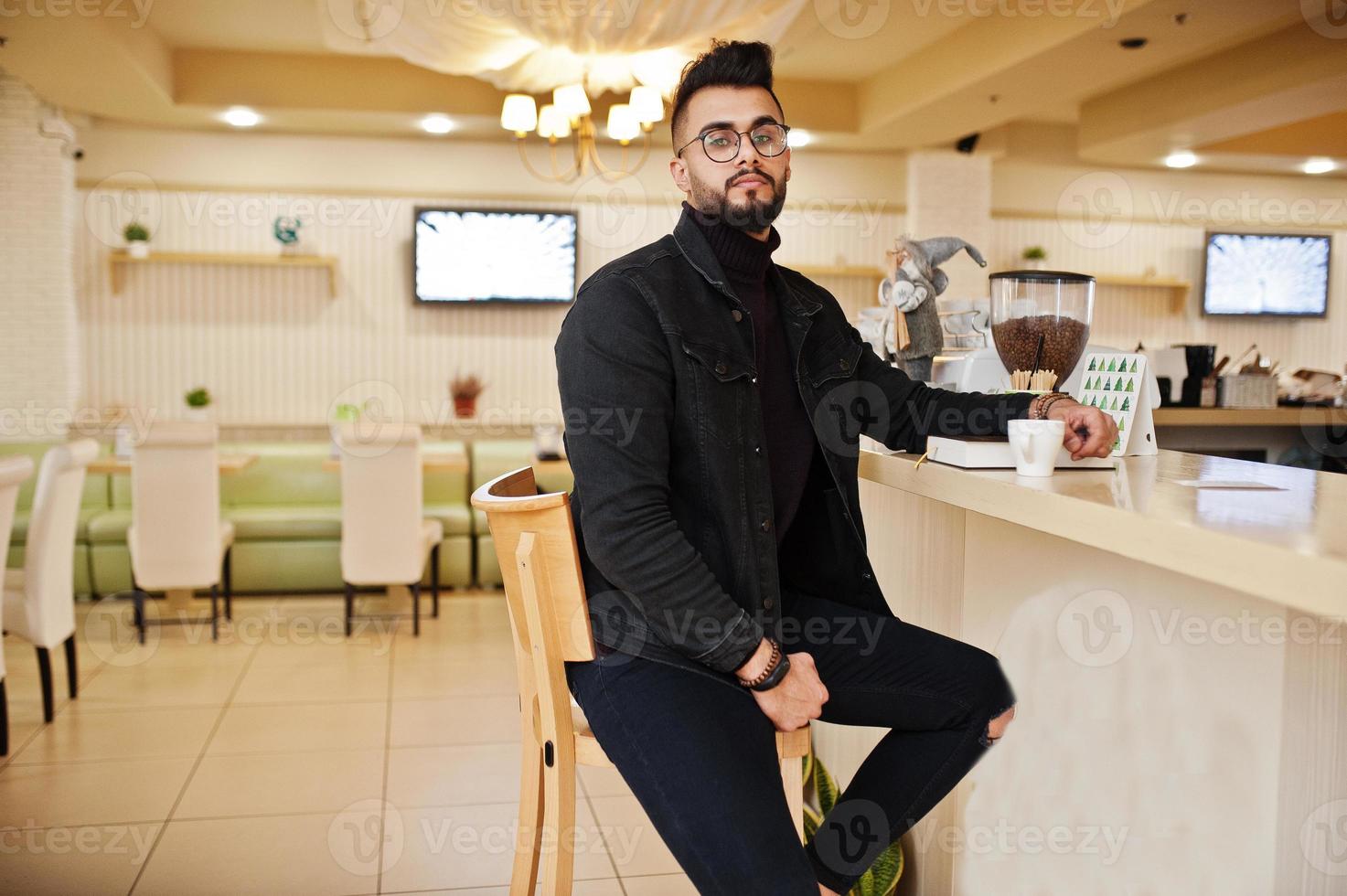 arabischer mann trägt schwarze jeansjacke und brille im café trinken kaffee an der bar mit buch. stilvoller und modischer arabischer Modelltyp. foto