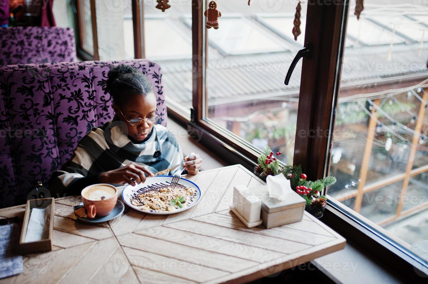 afrikanische frau in kariertem umhang und brille, die im café sitzt und dessert isst. foto