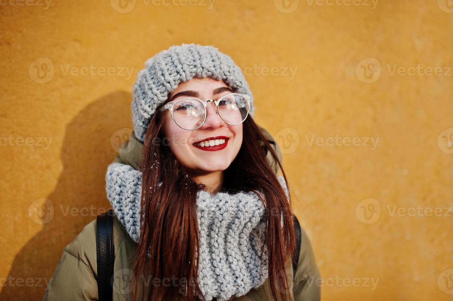 Porträt eines brünetten Mädchens in grauem Schal und Hut, Brille bei kaltem Wetter mit Sonnenschein gegen orangefarbene Wand des alten Hauses. freier Kopierplatz. foto
