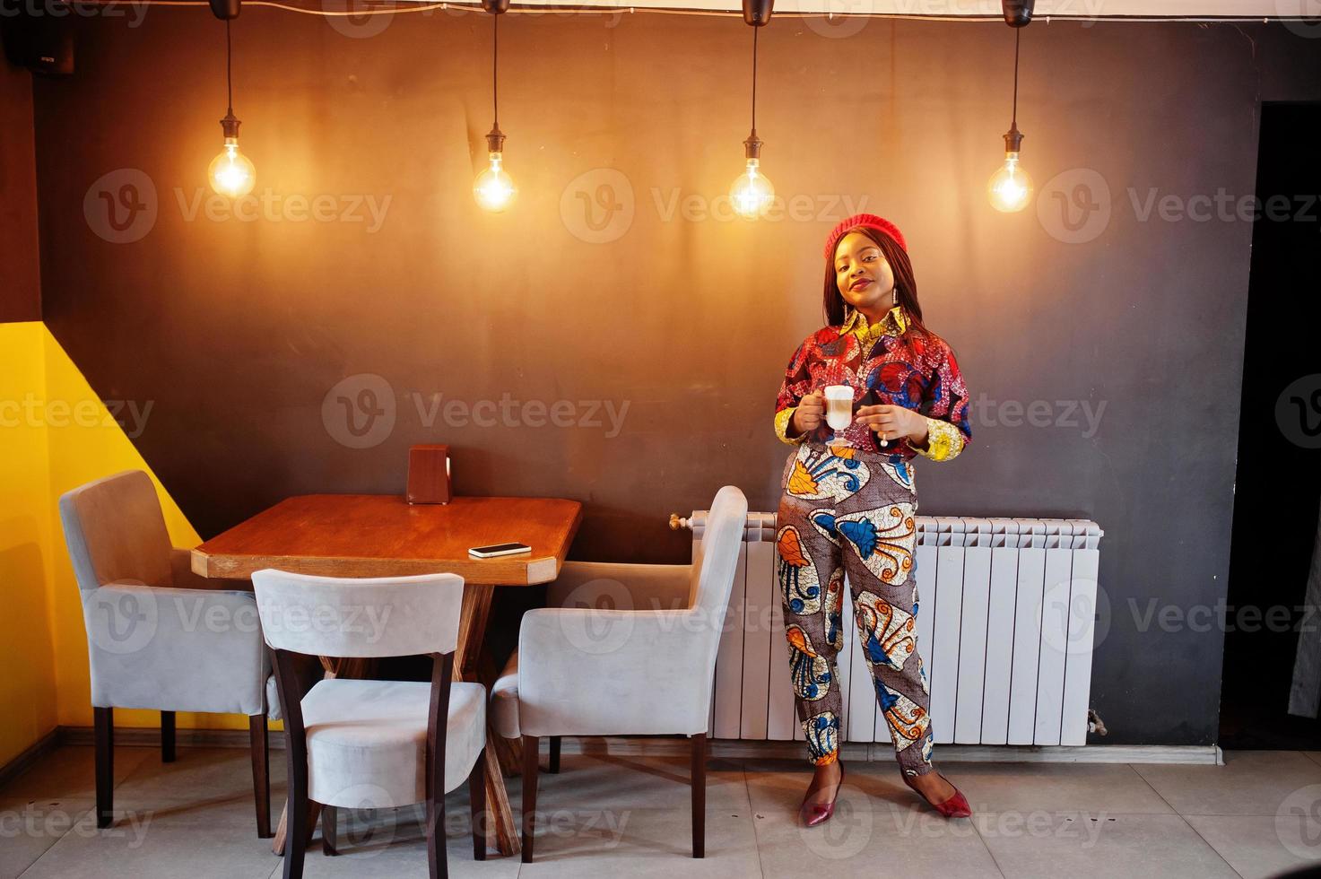 begeisterte afroamerikanische frau im trendigen farbigen outfit mit roter mütze, die in einem gemütlichen café mit einer tasse heißem latte in den händen chillt. foto