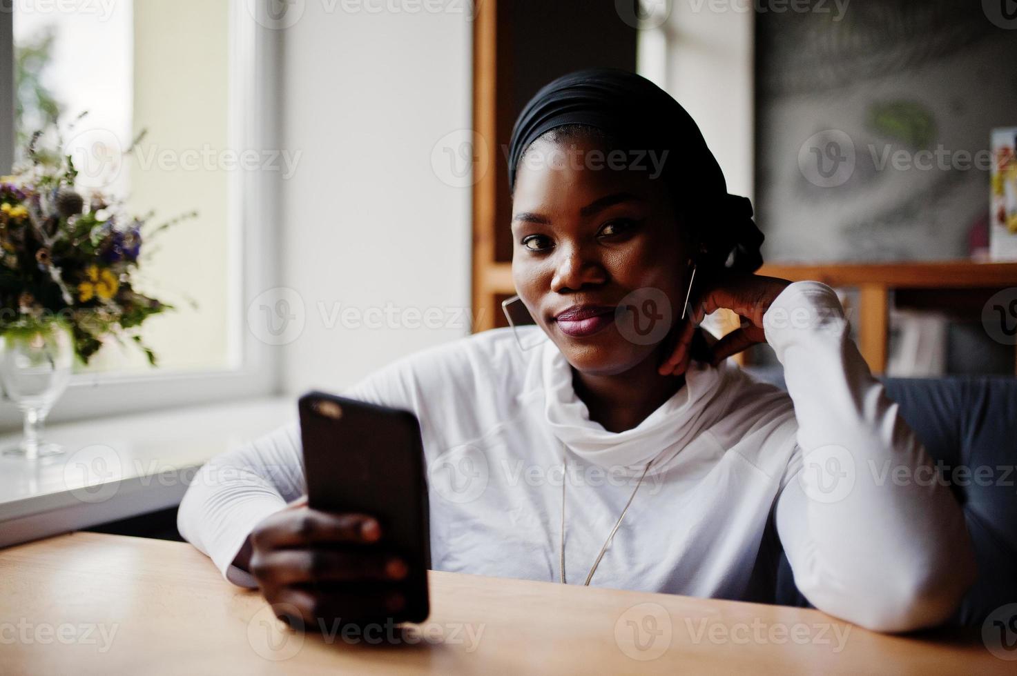 Afrikanisches muslimisches Mädchen in schwarzem Hijab sitzt im Café mit Handy zur Hand. foto