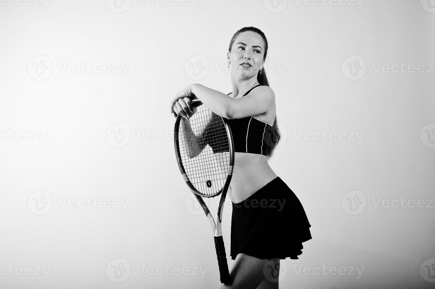 Schwarz-Weiß-Porträt einer schönen jungen Spielerin in Sportkleidung, die einen Tennisschläger hält, während sie vor weißem Hintergrund steht. foto