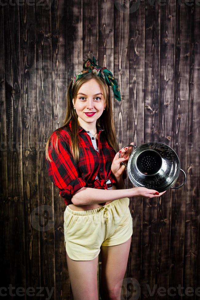 junge lustige hausfrau in kariertem hemd und gelben shorts pin-up-stil mit küchensieb auf hölzernem hintergrund. foto