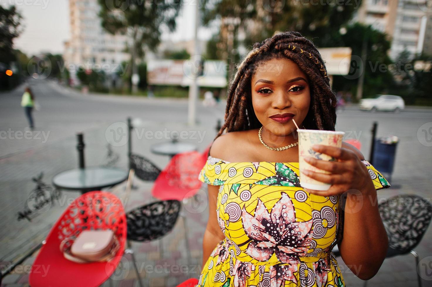 süßes kleines afrikanisch-amerikanisches Mädchen mit Dreadlocks, trägt ein farbiges gelbes Kleid, posiert im Café im Freien mit einer Tasse Kaffee. foto