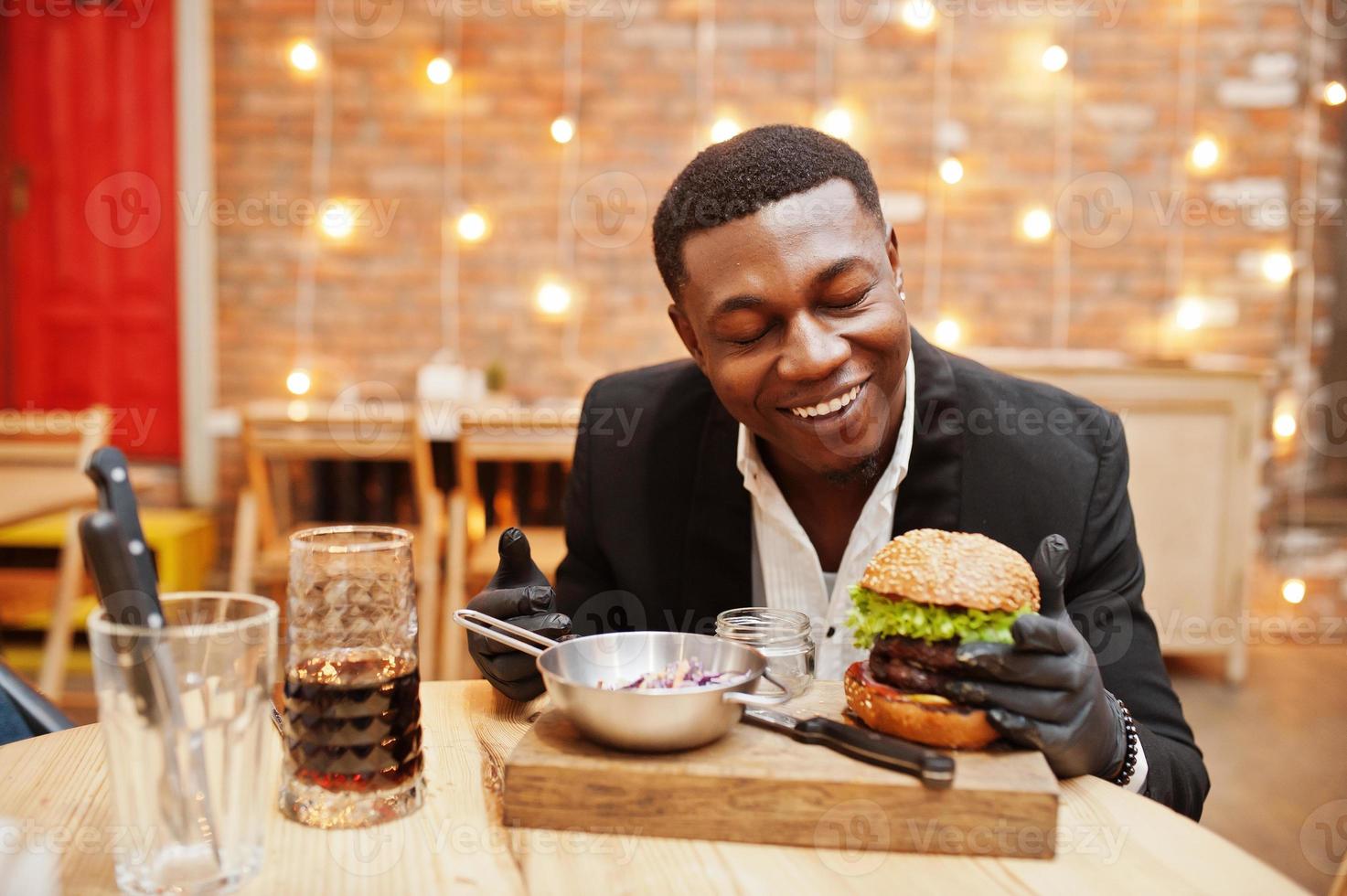 respektabler junger Afroamerikaner in schwarzem Anzug und Handschuhen zum Essen, der im Restaurant mit leckerem Doppelburger und Sodagetränk sitzt. foto