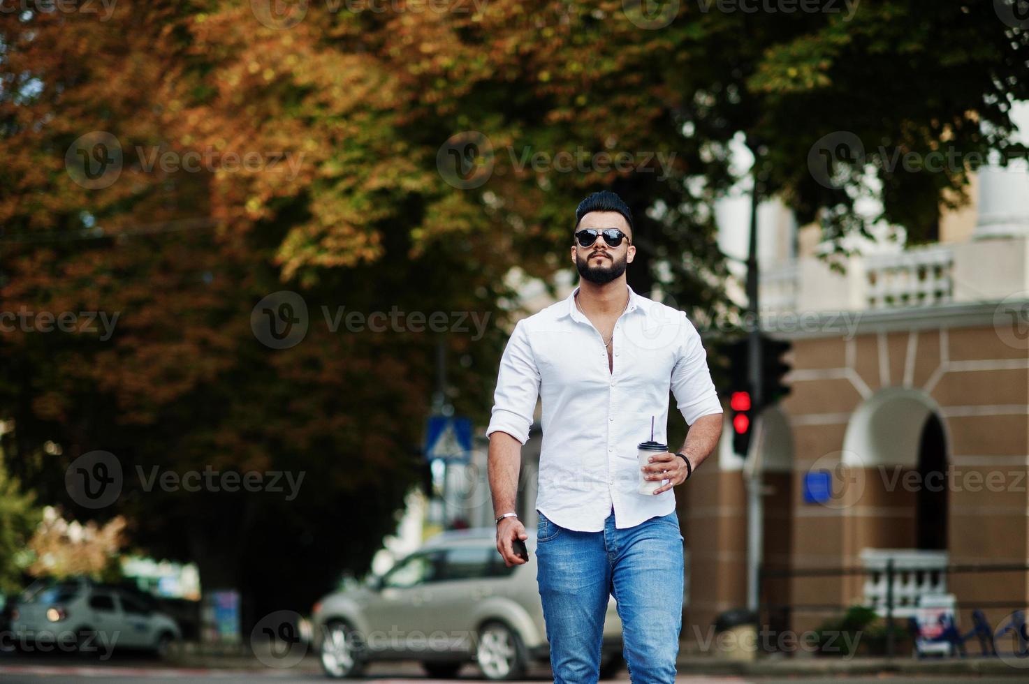 Stilvolles großes arabisches Mannmodell in weißem Hemd, Jeans und Sonnenbrille posierte auf der Straße der Stadt. bart attraktiver arabischer kerl mit tasse kaffee zu fuß. foto