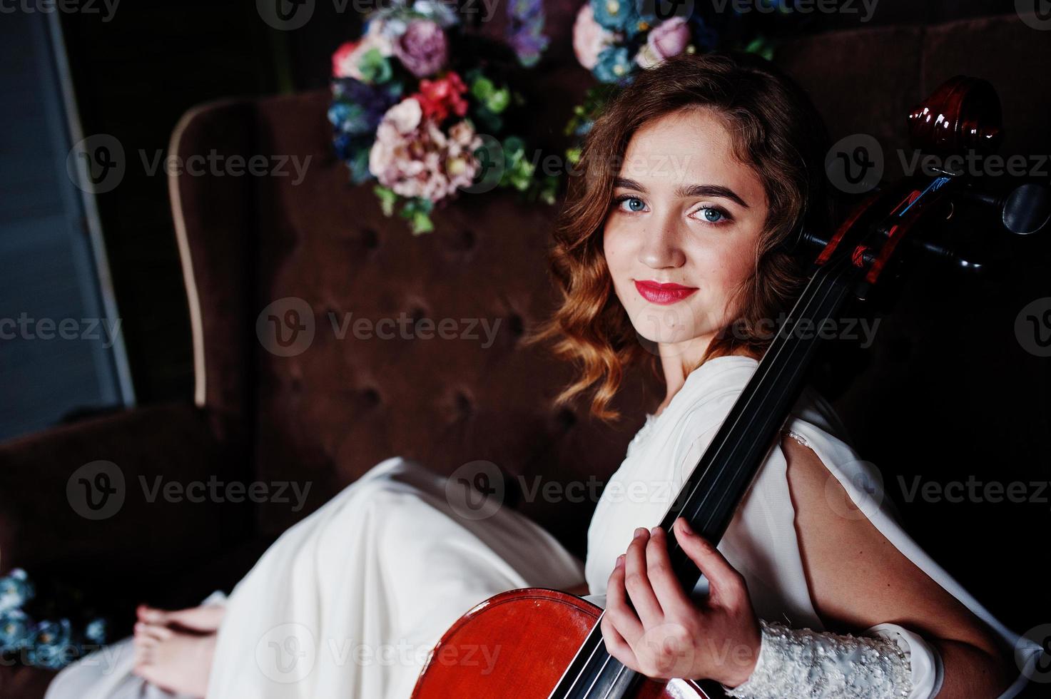 hübscher junger Mädchenmusiker im weißen Kleid mit Kontrabass, der auf braunem Vintage-Sofa sitzt. foto