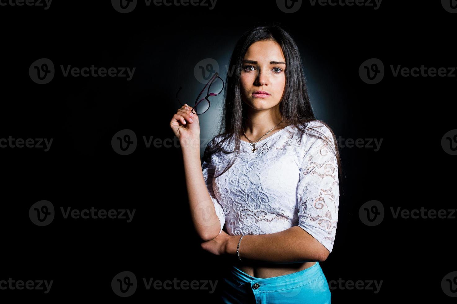 Porträt einer attraktiven jungen Frau in weißem Oberteil und blauer Hose, die mit ihrer Brille im Dunkeln posiert. foto