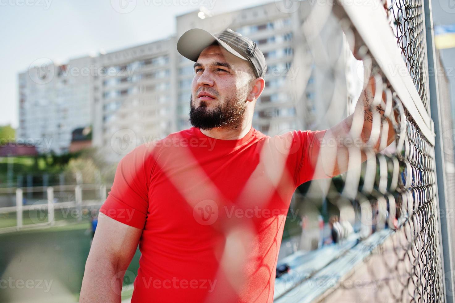 junger brutaler bärtiger muskulöser mann trägt rotes hemd, shorts und mütze im stadion. foto