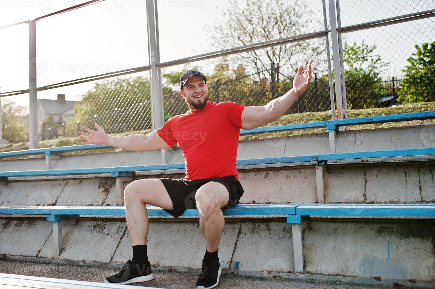 junger brutaler bärtiger muskulöser mann trägt rotes hemd, shorts und mütze im stadion. foto