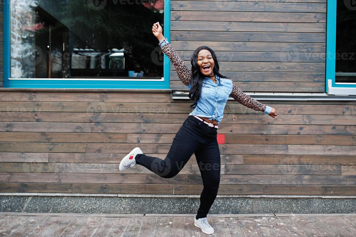Hipster afroamerikanische Mädchen mit Jeanshemd mit Leopardenärmeln springen auf der Straße gegen Holzhaus mit Fenstern. foto