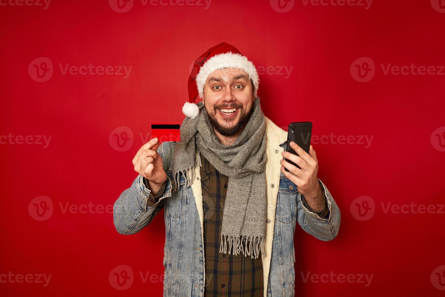 Aufgeregter Mann mit Weihnachtsmütze hält Bankkreditkarte und kauft Telefon über mobile Anwendung im Online-Shop Geschenke für das neue Jahr isoliertes rotes Studio-Hintergrundkonzept - Menschen, Technologie, Einkaufen foto