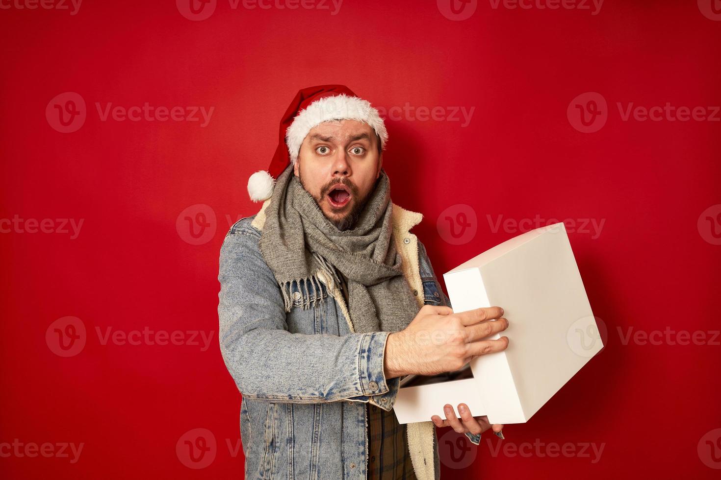 überraschter aufgeregter mann mit weihnachtsmütze, jeansjacke öffnet weiße schachtel, die sie in seinen händen hält, schockiert über geschenk für neujahr. isolierter roter Studiohintergrund. Konzept - Urlaub, Emotionen, Menschen foto