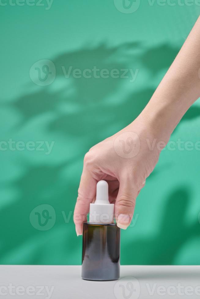weibliche Hand hält kosmetisches Produkt in Tube, Glasflasche, Lotion, Anti-Aging-Feuchtigkeitscreme, ätherisches Gesichtsöl oder Serum auf grünem Hintergrund mit heller Schattennatur. kosmetisches hautpflegekonzept der schönheit. foto