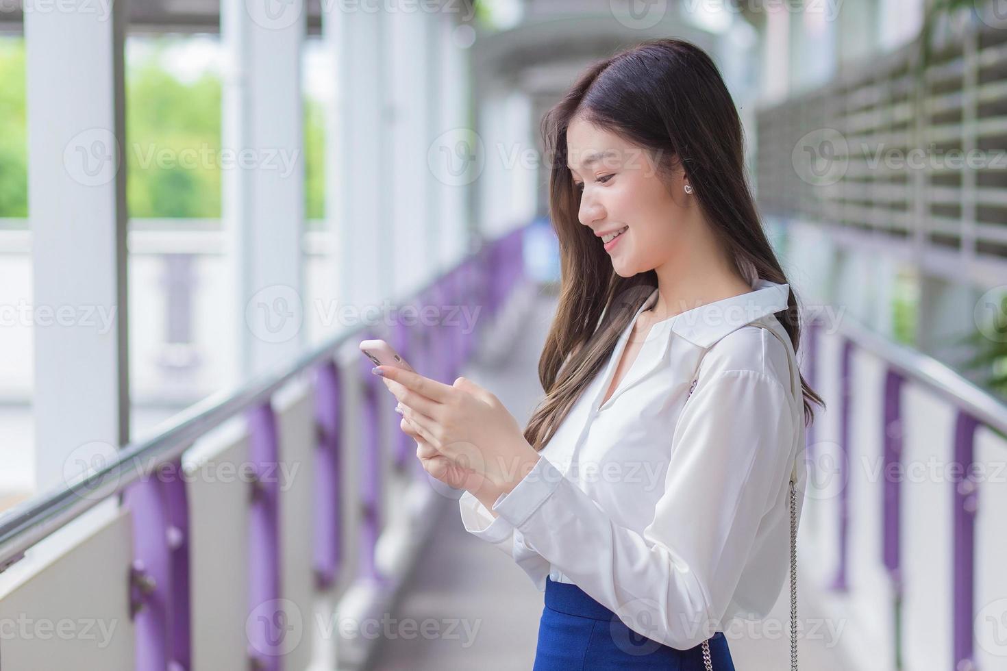 Professionelle, schöne, selbstbewusste junge asiatische Geschäftsfrau steht auf der Überführung des Skytrain im Freien in der Stadt, während sie ihr Smartphone verwendet, um Nachrichten an das Büro zu senden. foto