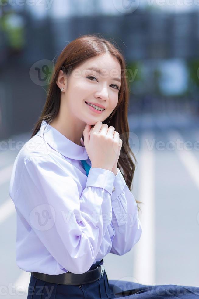 Schönes asiatisches Gymnasiastmädchen in der Schuluniform mit Zahnspangen sitzt und lächelt selbstbewusst, während sie glücklich mit dem Gebäude im Hintergrund in die Kamera schaut. foto