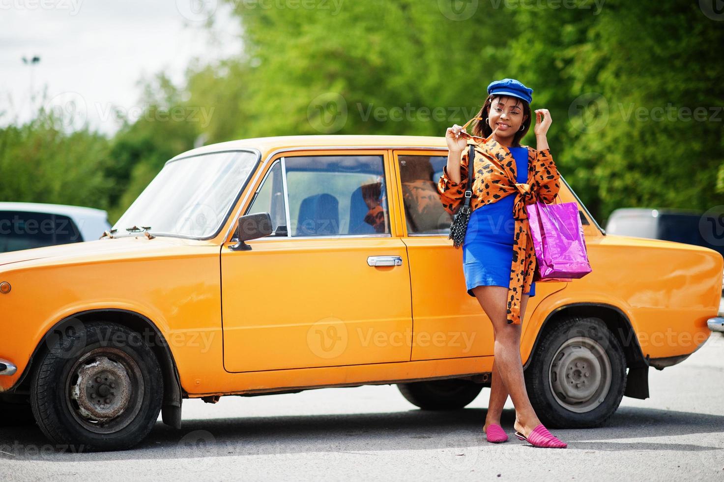 Schöne afrikanisch-amerikanische Dame mit Einkaufstüten, die in der Nähe eines orangefarbenen klassischen Retro-Autos steht. foto