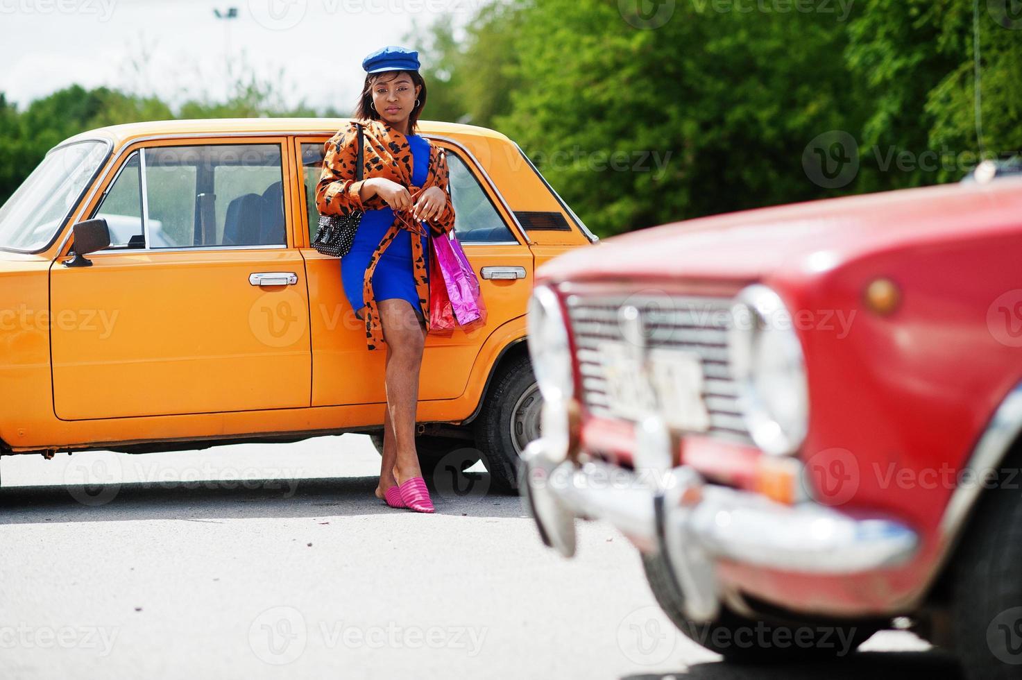 Schöne afrikanisch-amerikanische Dame mit Einkaufstüten, die in der Nähe eines orangefarbenen klassischen Retro-Autos steht. foto