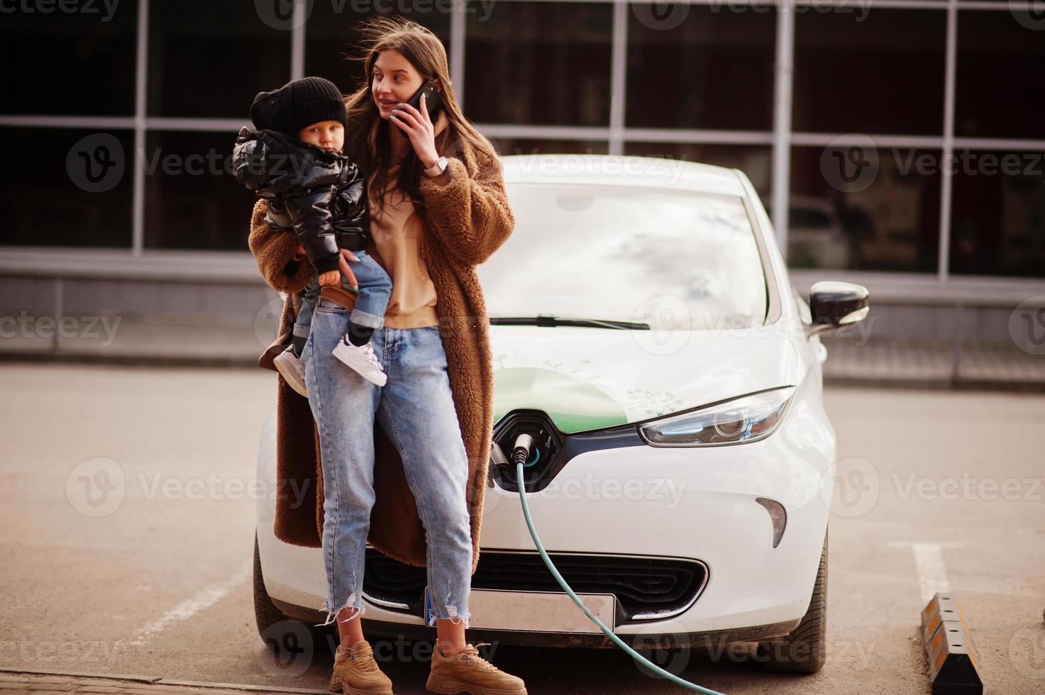 junge mutter mit kind, das elektroauto an der elektrotankstelle auflädt und am handy spricht. foto