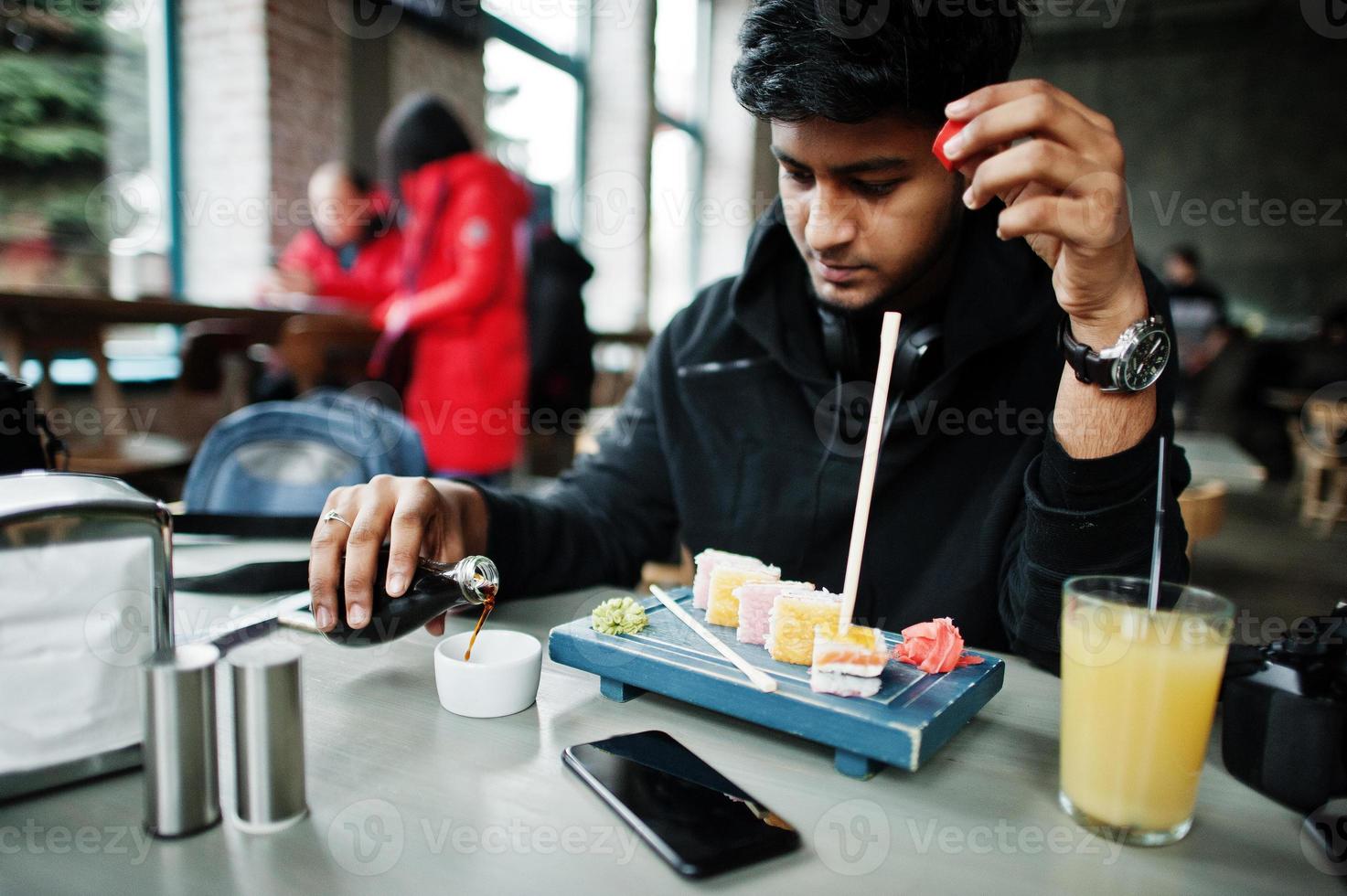 Lässiger und stilvoller junger asiatischer Mann mit Kopfhörern im Café, der Sushi isst und Sojasauce gießt. foto