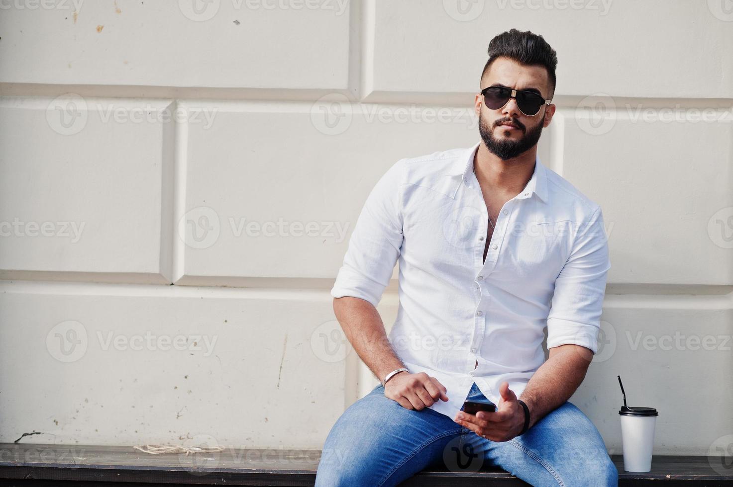 Stilvolles großes arabisches Mannmodell in weißem Hemd, Jeans und Sonnenbrille posiert auf der Straße der Stadt und hält das Handy zur Hand. bart arabischer kerl mit einer tasse kaffee sitzen und sich ausruhen. foto