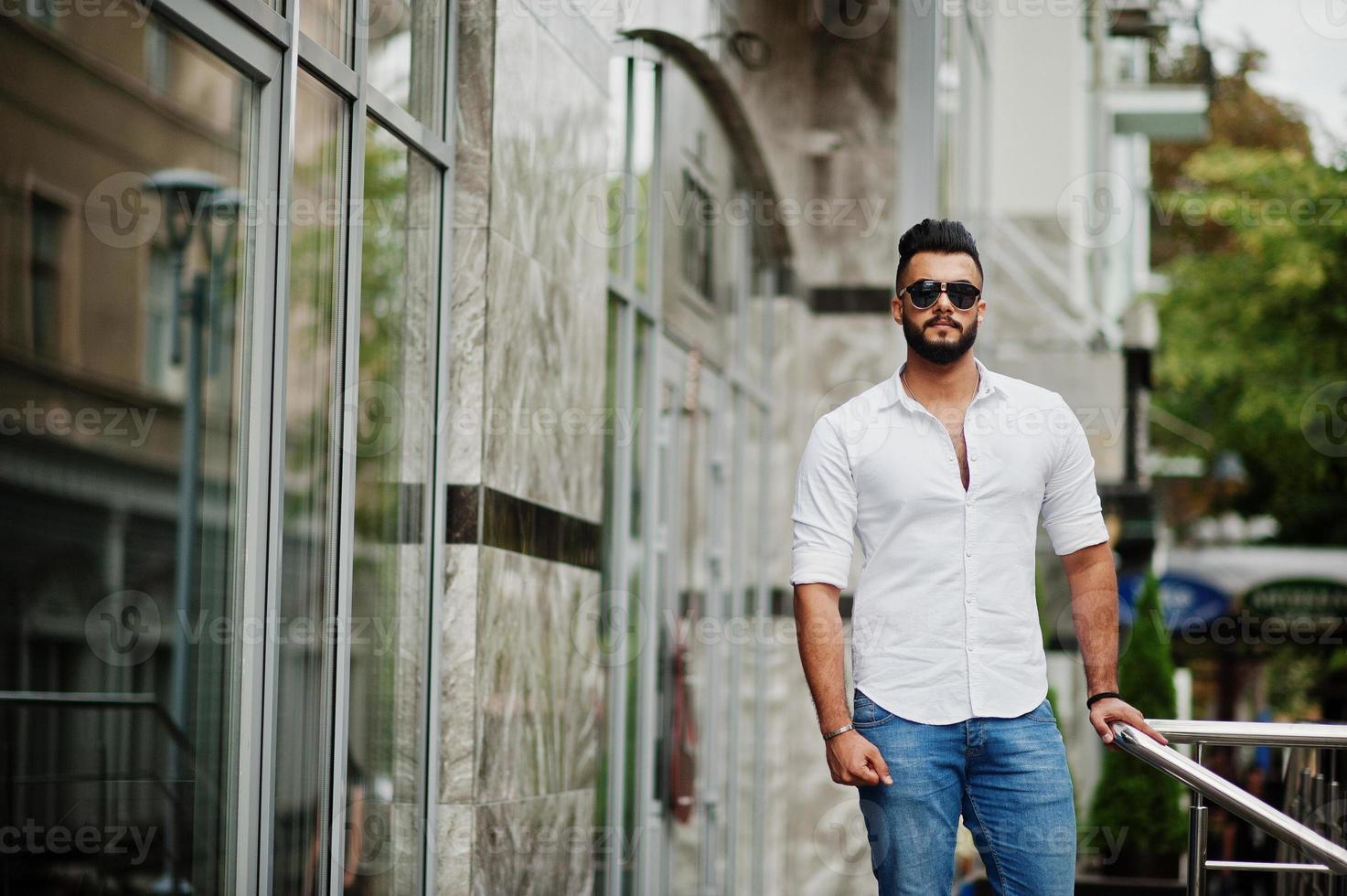 Stilvolles großes arabisches Mannmodell in weißem Hemd, Jeans und Sonnenbrille posierte auf der Straße der Stadt. Bart attraktiver arabischer Typ. foto