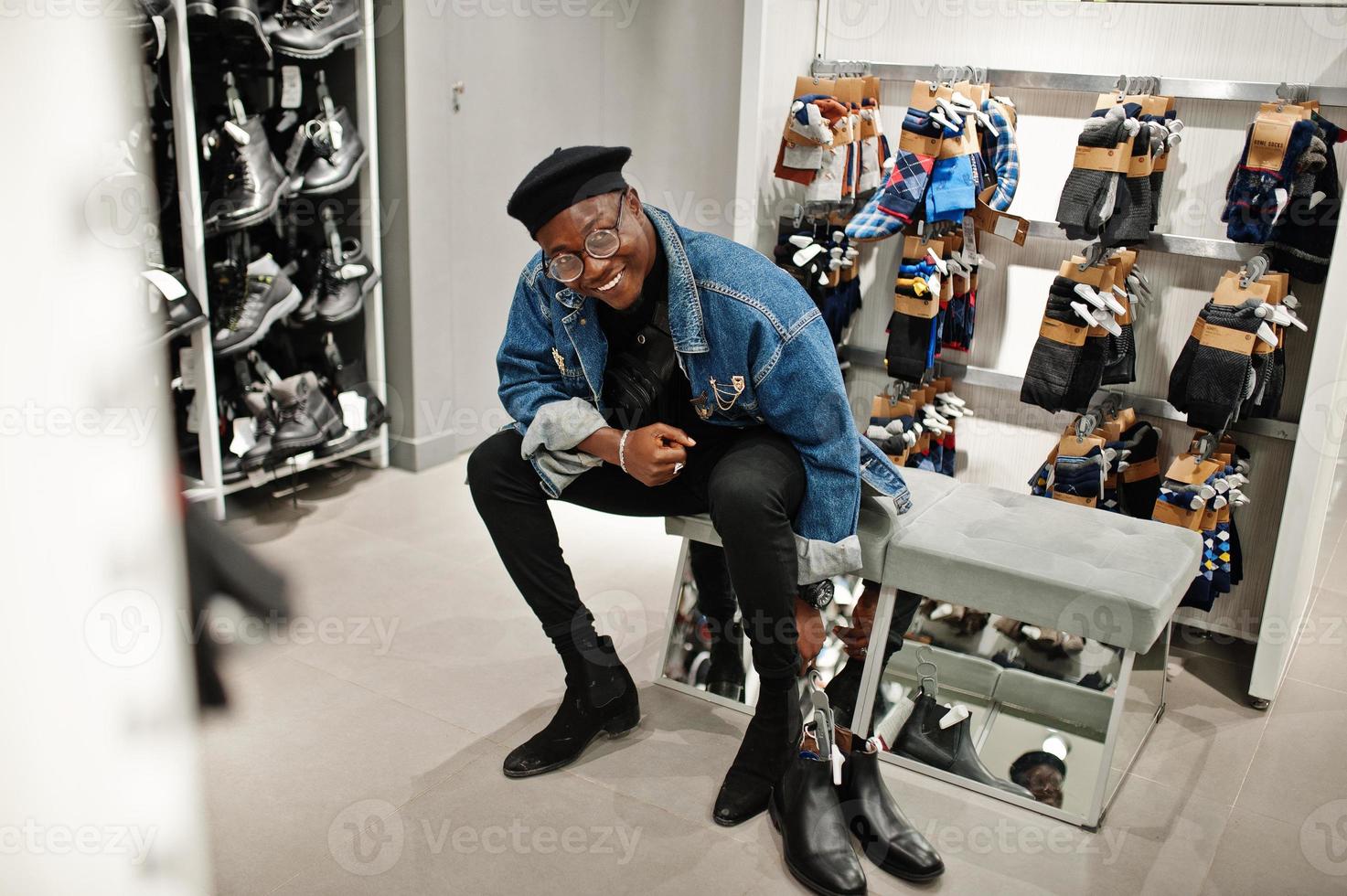 stilvoller, lässiger afroamerikanischer Mann in Jeansjacke und schwarzer Baskenmütze im Bekleidungsgeschäft, der neue Schuhe ausprobiert. foto