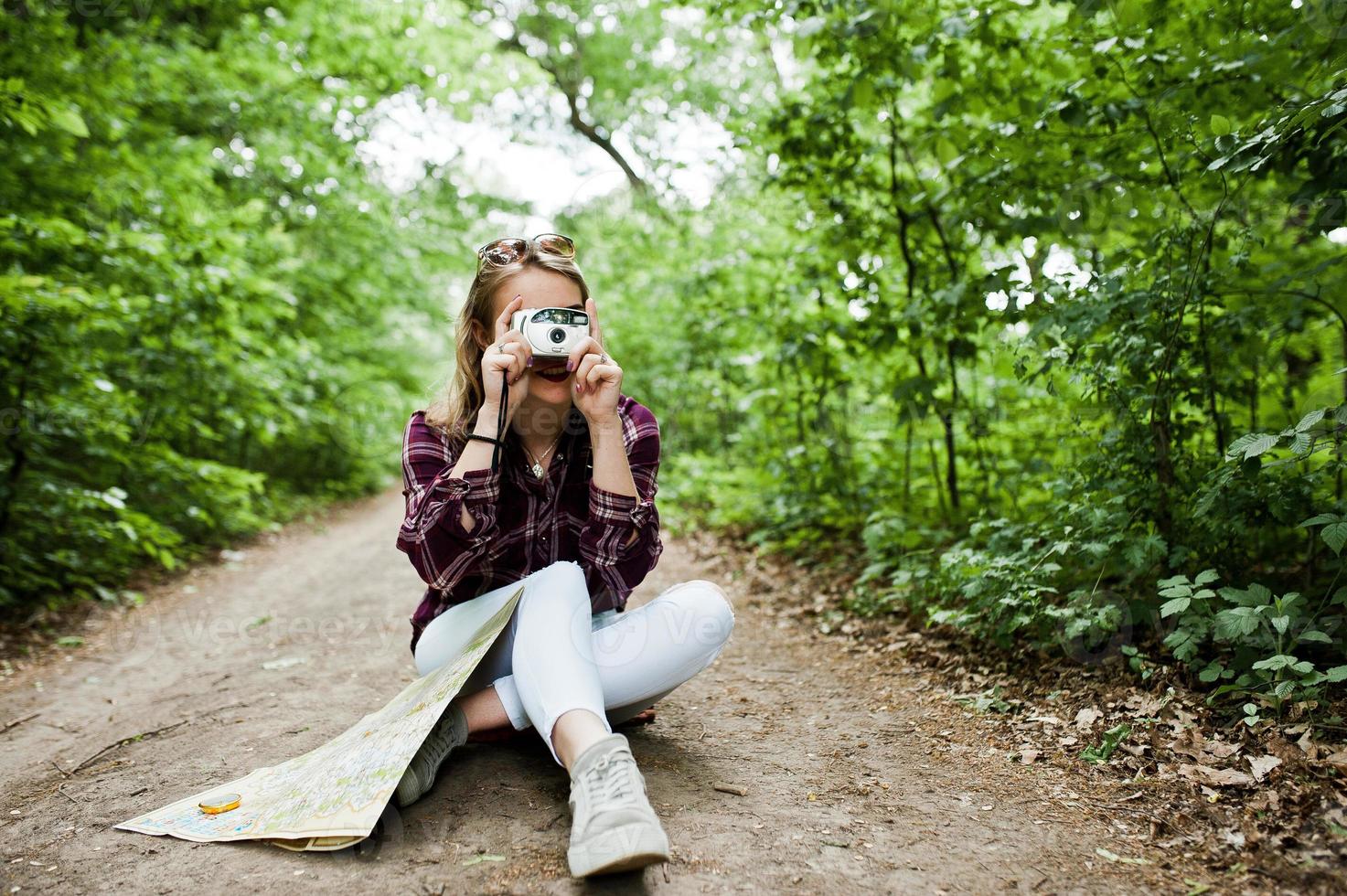 Porträt eines attraktiven blonden Mädchens mit einer Karte, das im Wald sitzt und Fotos macht.