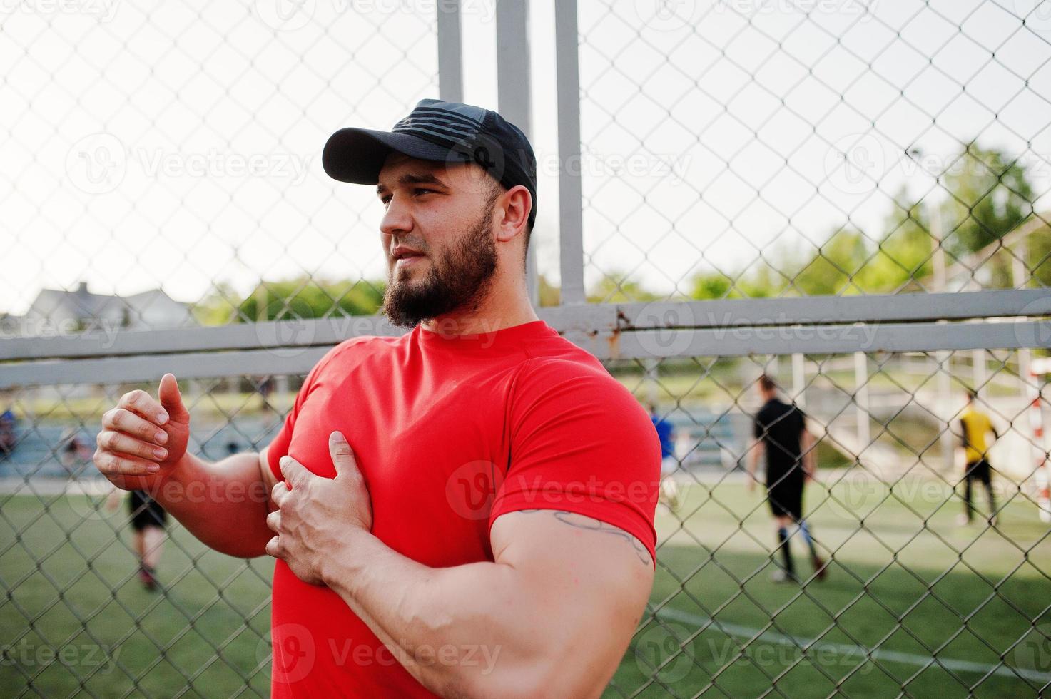 junger brutaler bärtiger muskulöser mann trägt rotes hemd, shorts und mütze im stadion. foto