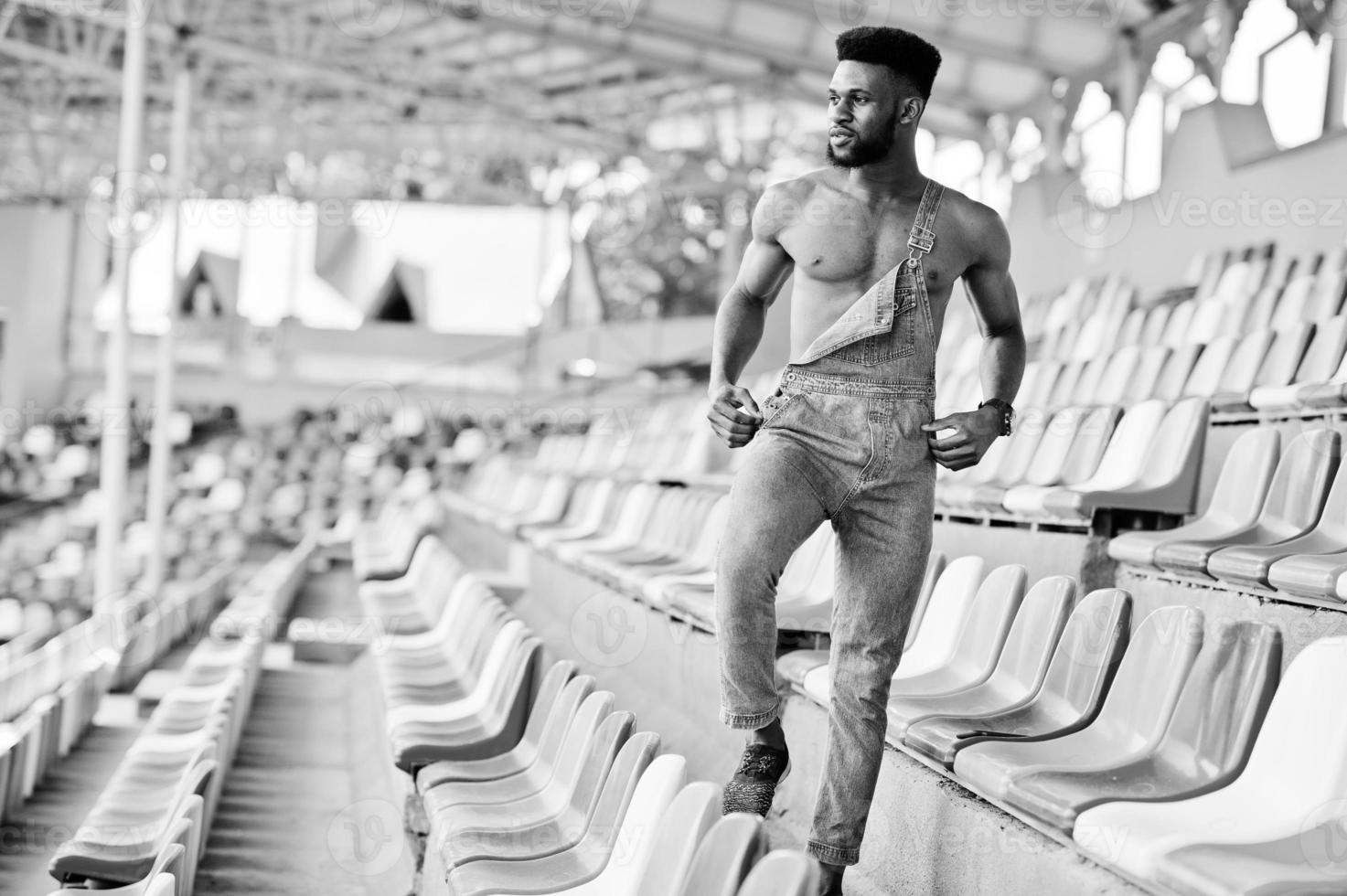 hübscher sexy afrikanisch-amerikanischer Mann mit nacktem Oberkörper in Jeans-Overalls posiert auf farbigen Stühlen im Stadion. modisches Porträt eines schwarzen Mannes. foto