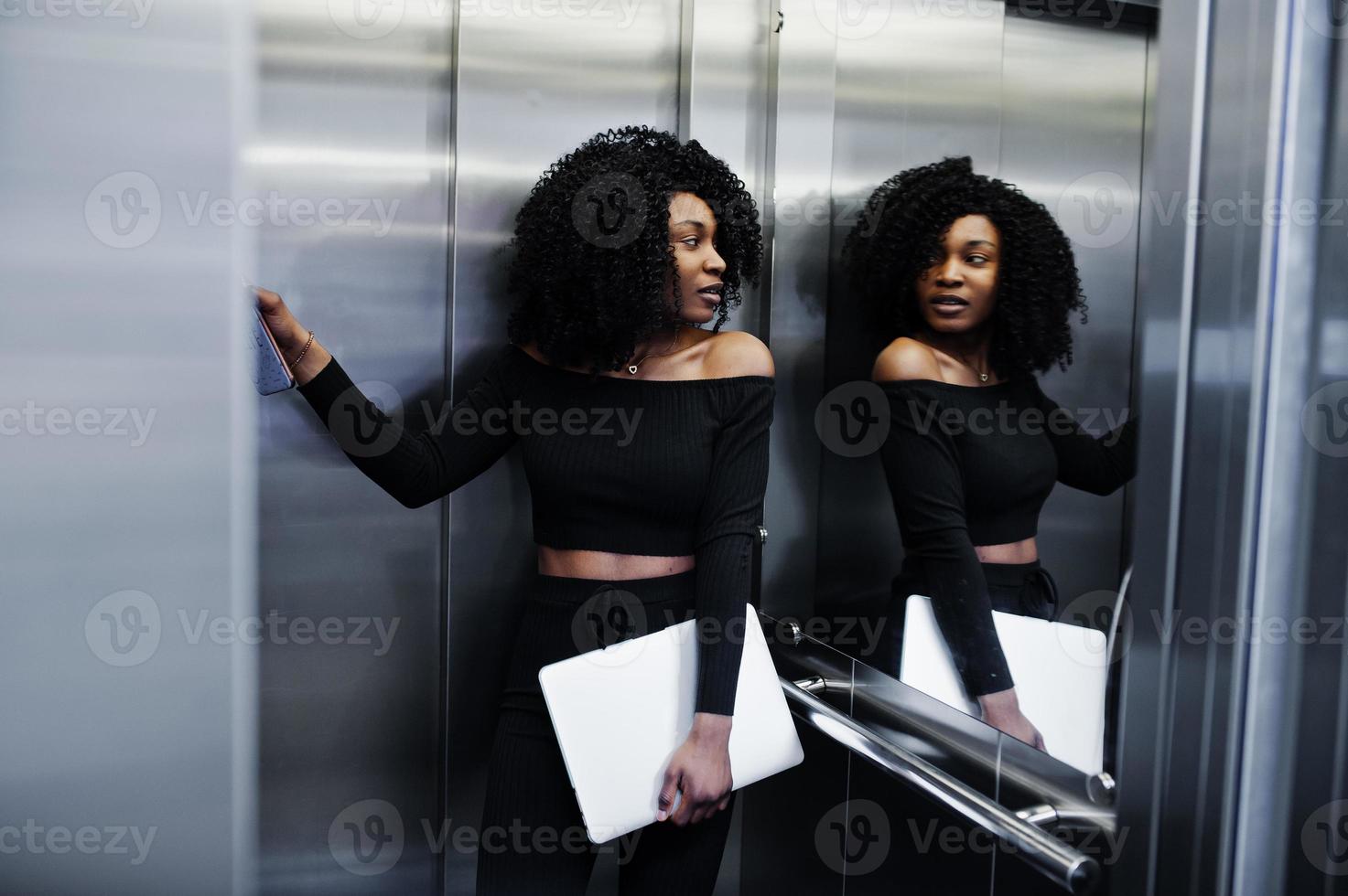 modische junge schöne afroamerikanische geschäftsfrau mit afrofrisur tragen in elegantem schwarz, stehen am aufzug mit laptop in der hand. foto