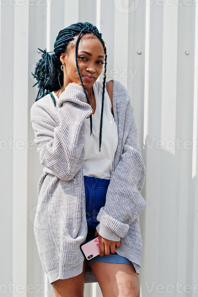 afrikanische frau mit dreadlockshaar, in jeansshorts, die mit handy in der hand vor einer weißen stahlwand posiert. foto