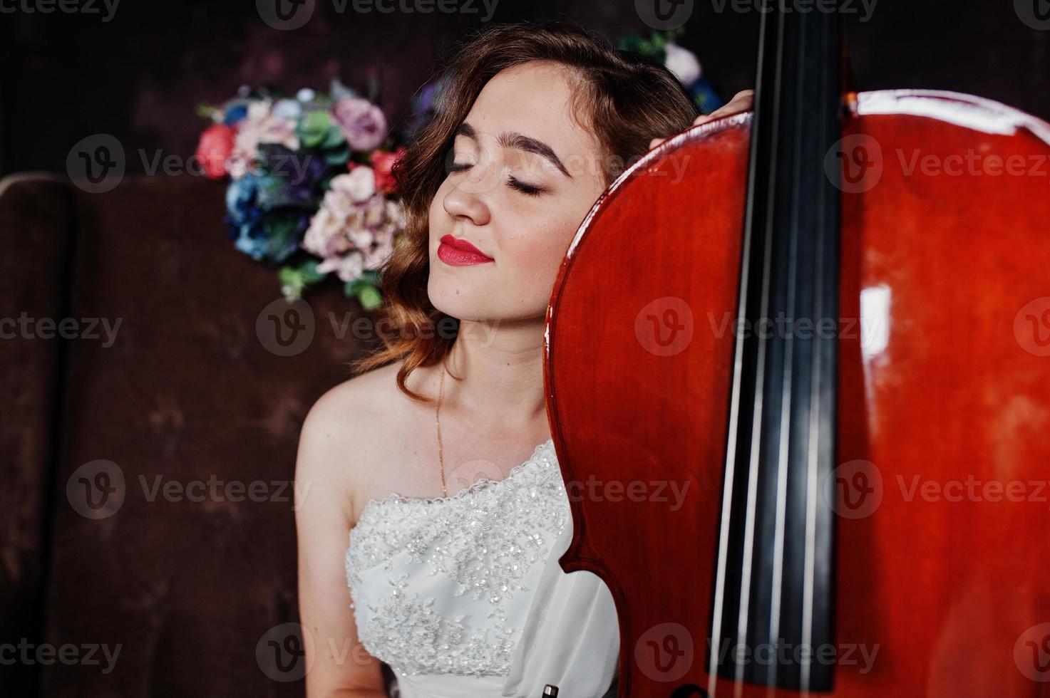 hübscher junger Mädchenmusiker im weißen Kleid mit Kontrabass, der auf braunem Vintage-Sofa sitzt. foto