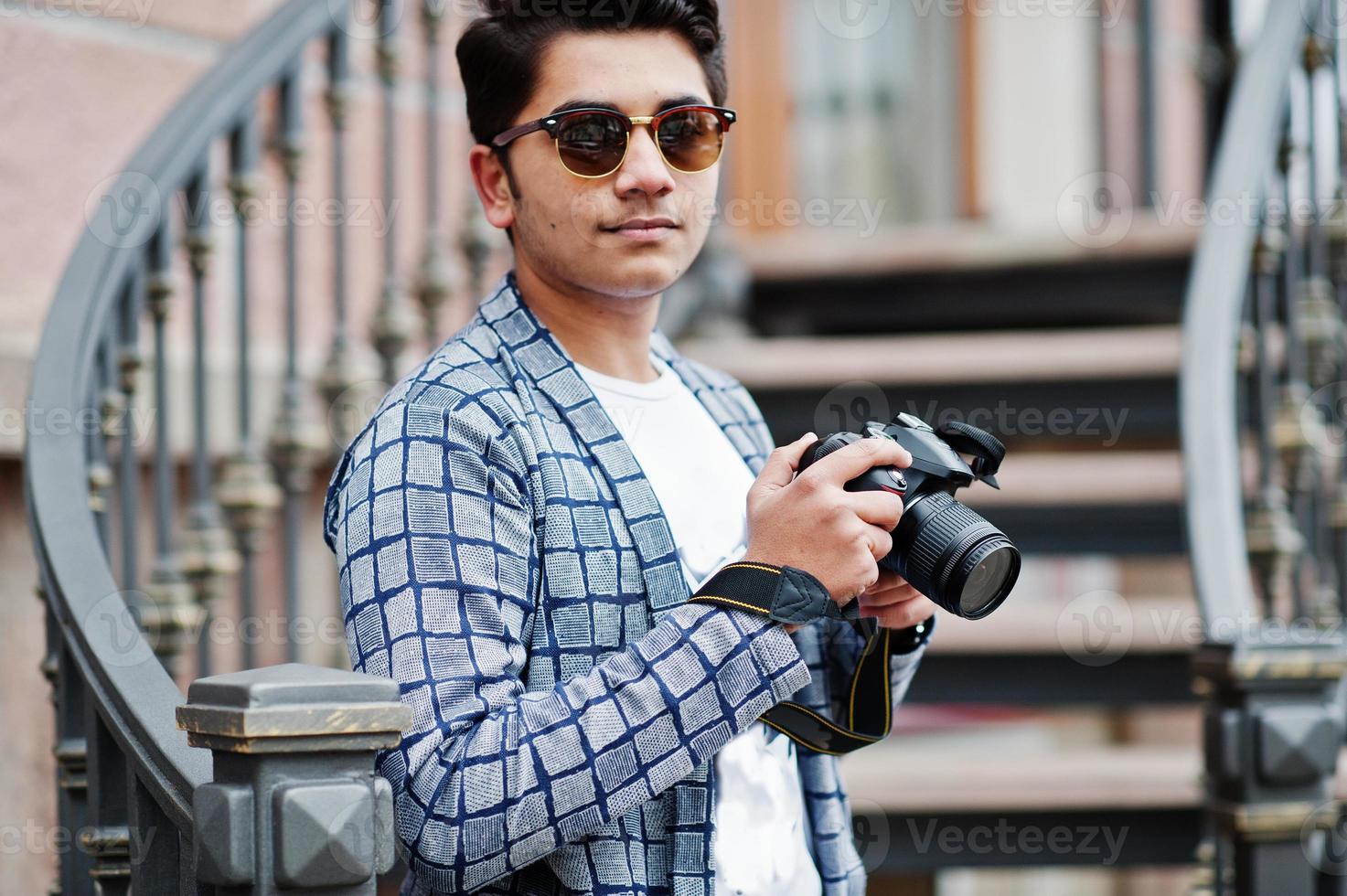 stilvoller indischer junger mannfotograf mit sonnenbrille trägt lässig posierte im freien gegen eisentreppe mit dslr-fotokamera an den händen. foto