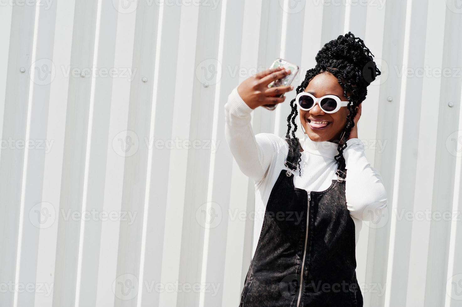 afrikanische frau im jeansoverallrock, weiße sonnenbrille posierte vor weißer stahlwand und machte selfie per telefon. foto