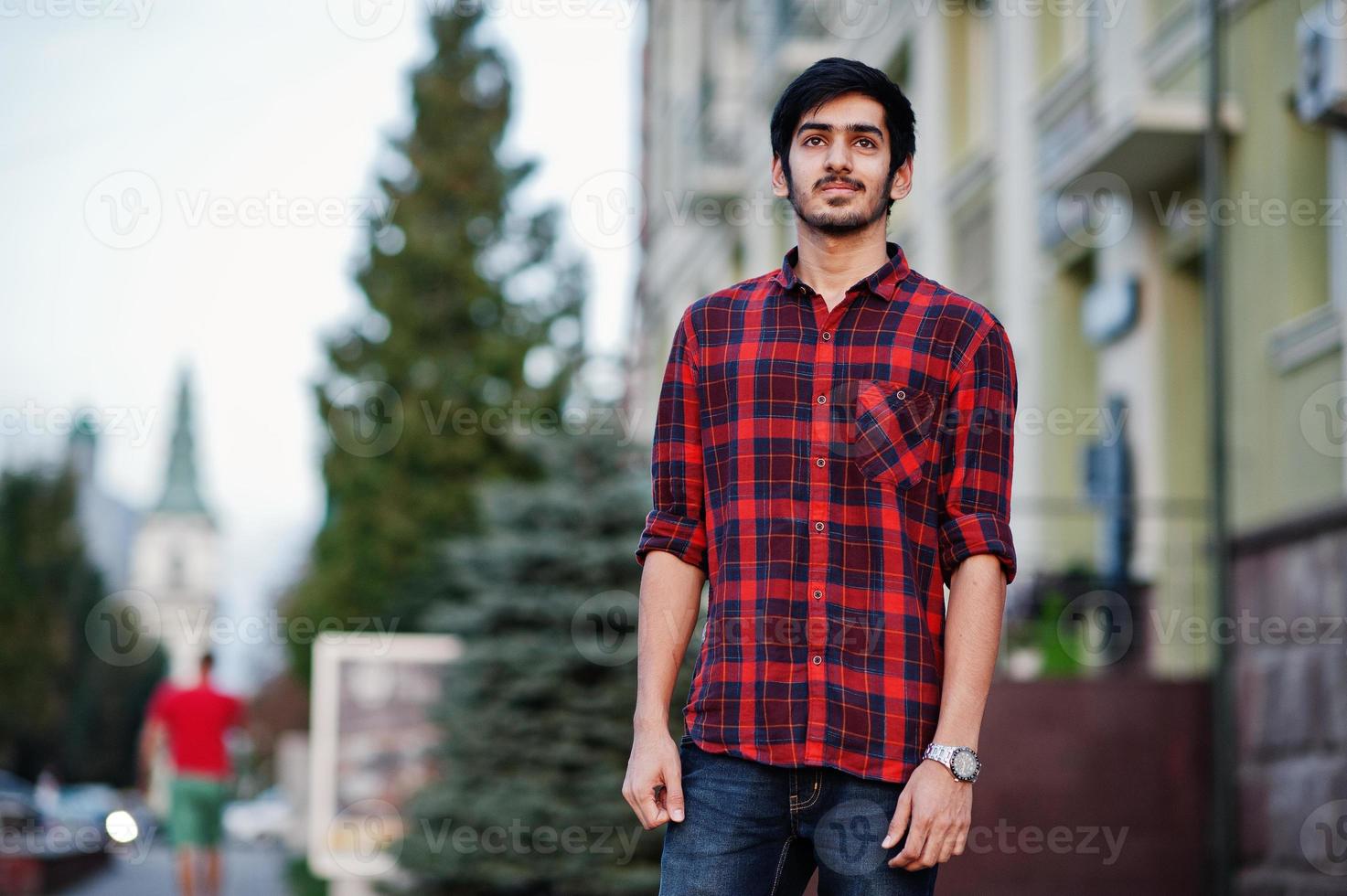 junger indischer studentenmann in rot kariertem hemd und jeans posierte in der stadt. foto