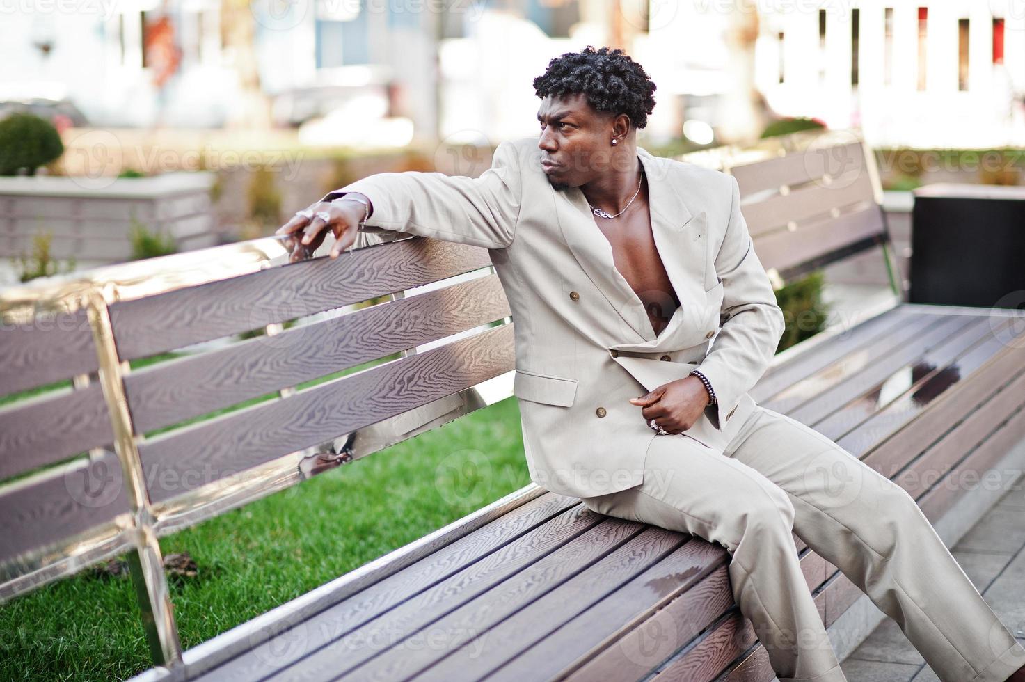 Stilvoller Afro-Mann im beigefarbenen Anzug der alten Schule sitzt auf der Bank. Modischer junger afrikanischer Mann in Freizeitjacke auf nacktem Oberkörper. foto