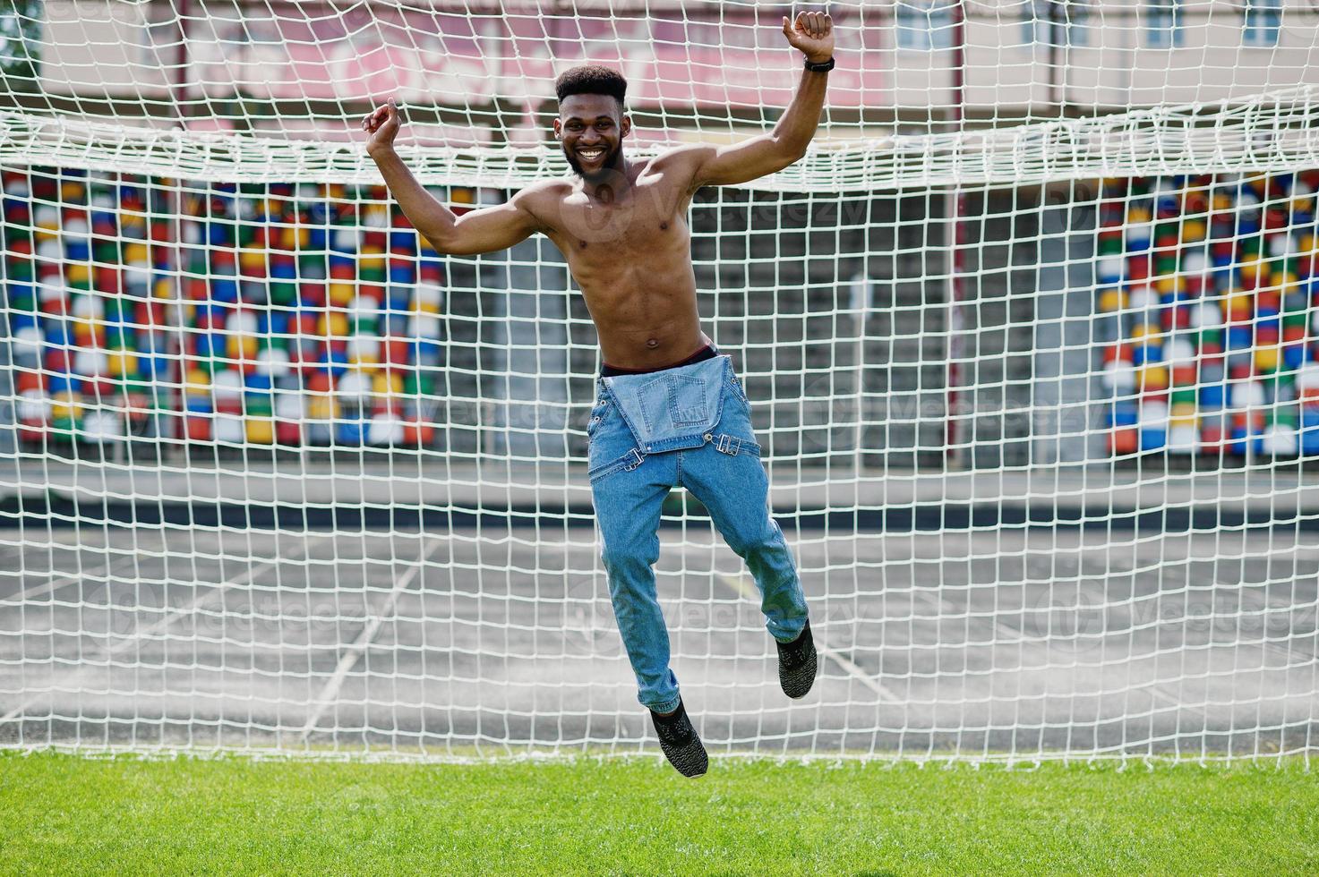 hübscher sexy afroamerikanischer muskulöser Mann mit nacktem Oberkörper in Jeans-Overalls posiert auf grünem Gras gegen Fußballtore des Stadionfeldes. modisches Porträt eines schwarzen Mannes. foto