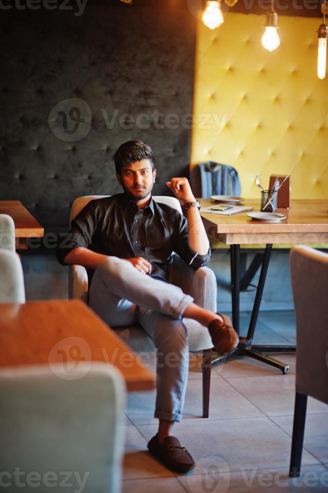 Selbstbewusster junger Inder im schwarzen Hemd, der im Café sitzt. foto